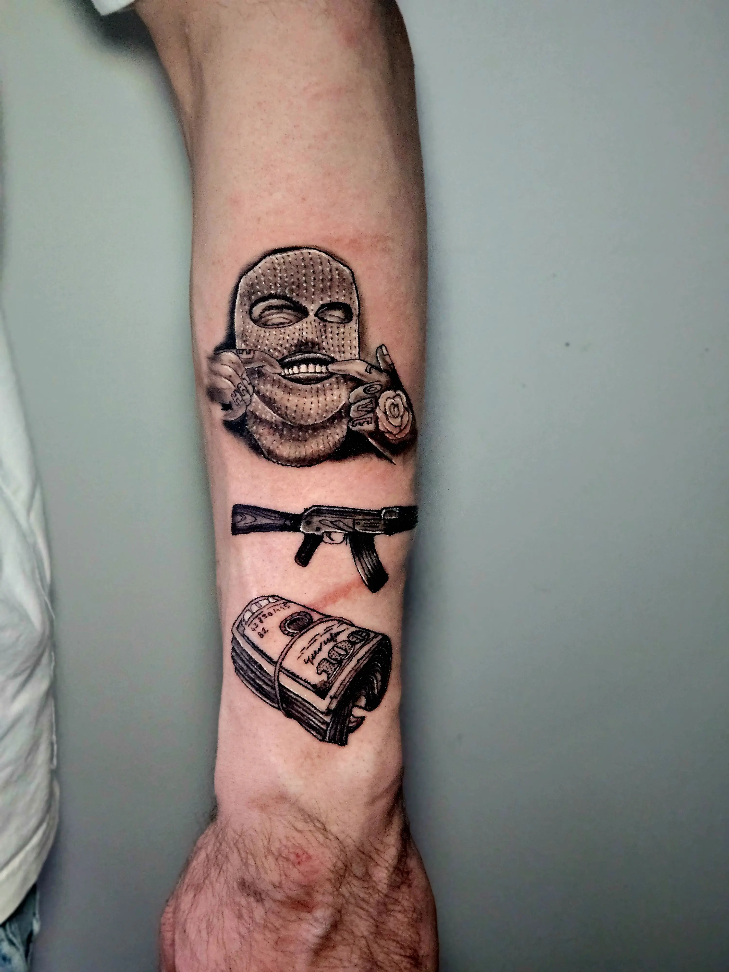 Tattoo depicting a masked figure, AK-47, and money roll in black and gray realism.