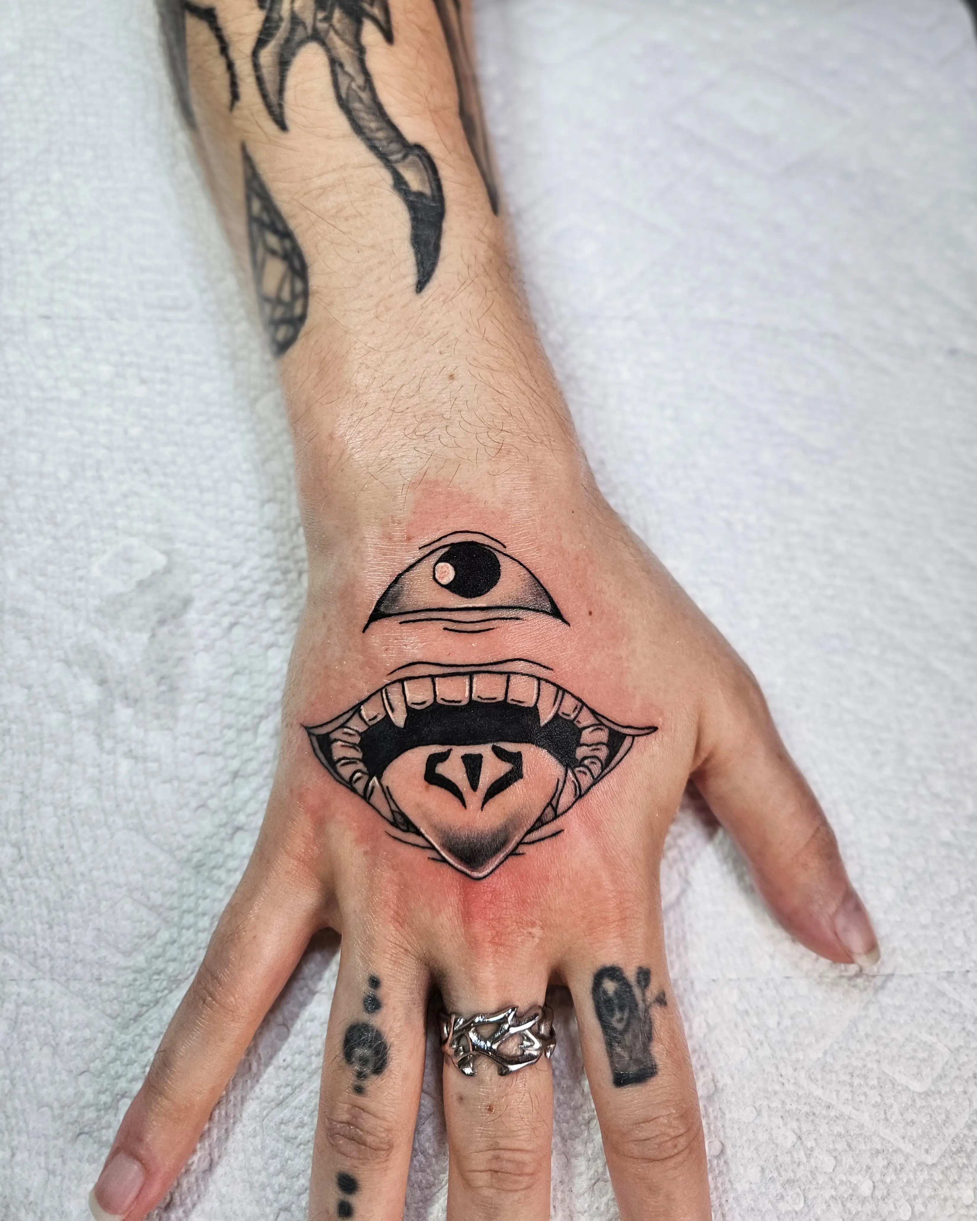 Surrealistic tattoo of an eye above a mouth with tongue detail on the hand.