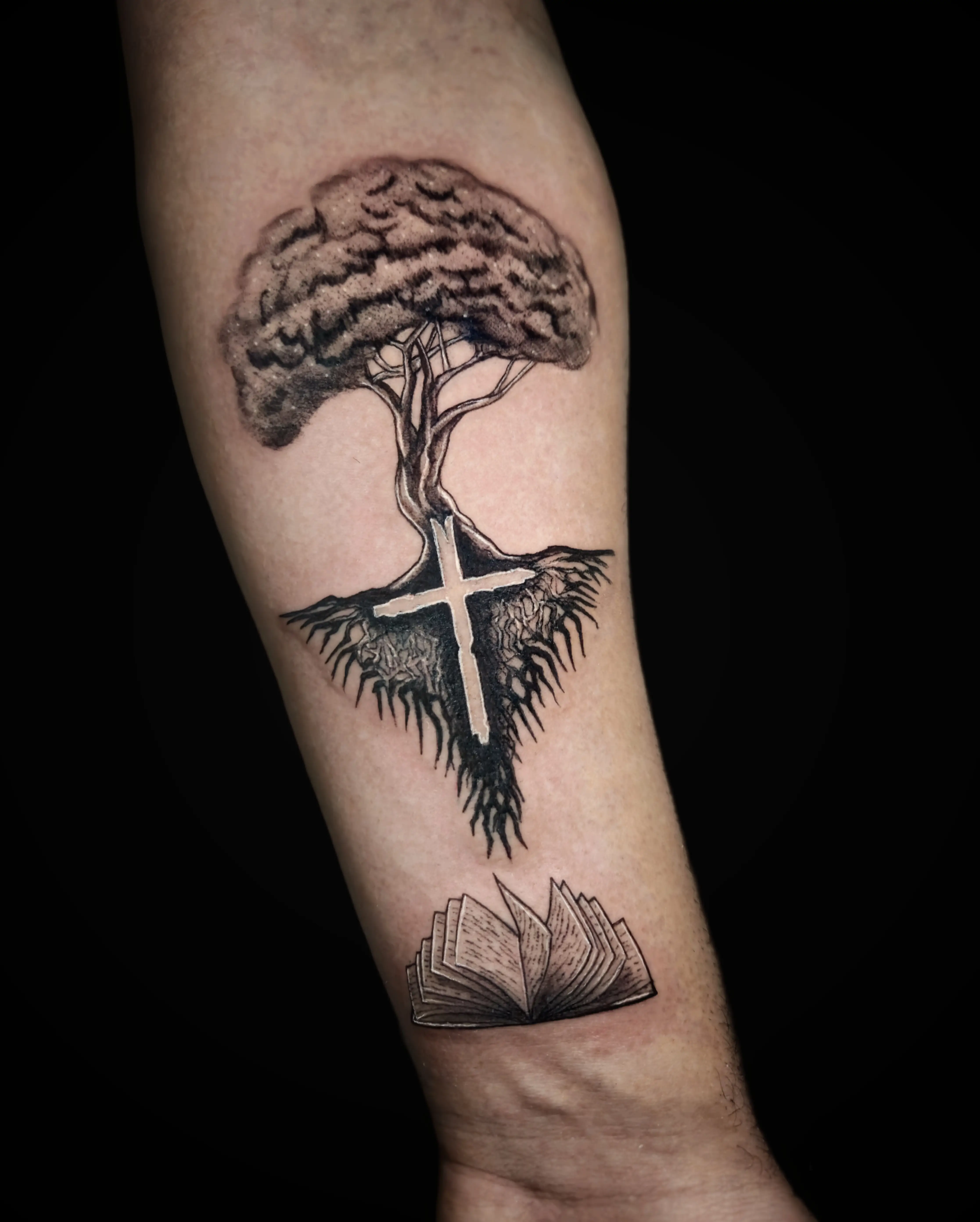 Tattoo of a tree with roots forming a cross and an open book beneath in black and gray style.