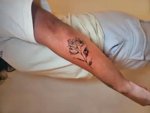 Delicate floral tattoo with a realistic red ladybug sitting on the stem, placed on the forearm.