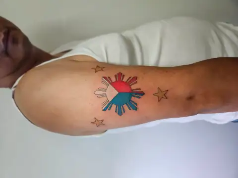 Philippine flag-inspired tattoo with sun and three stars in bold color on the upper arm.