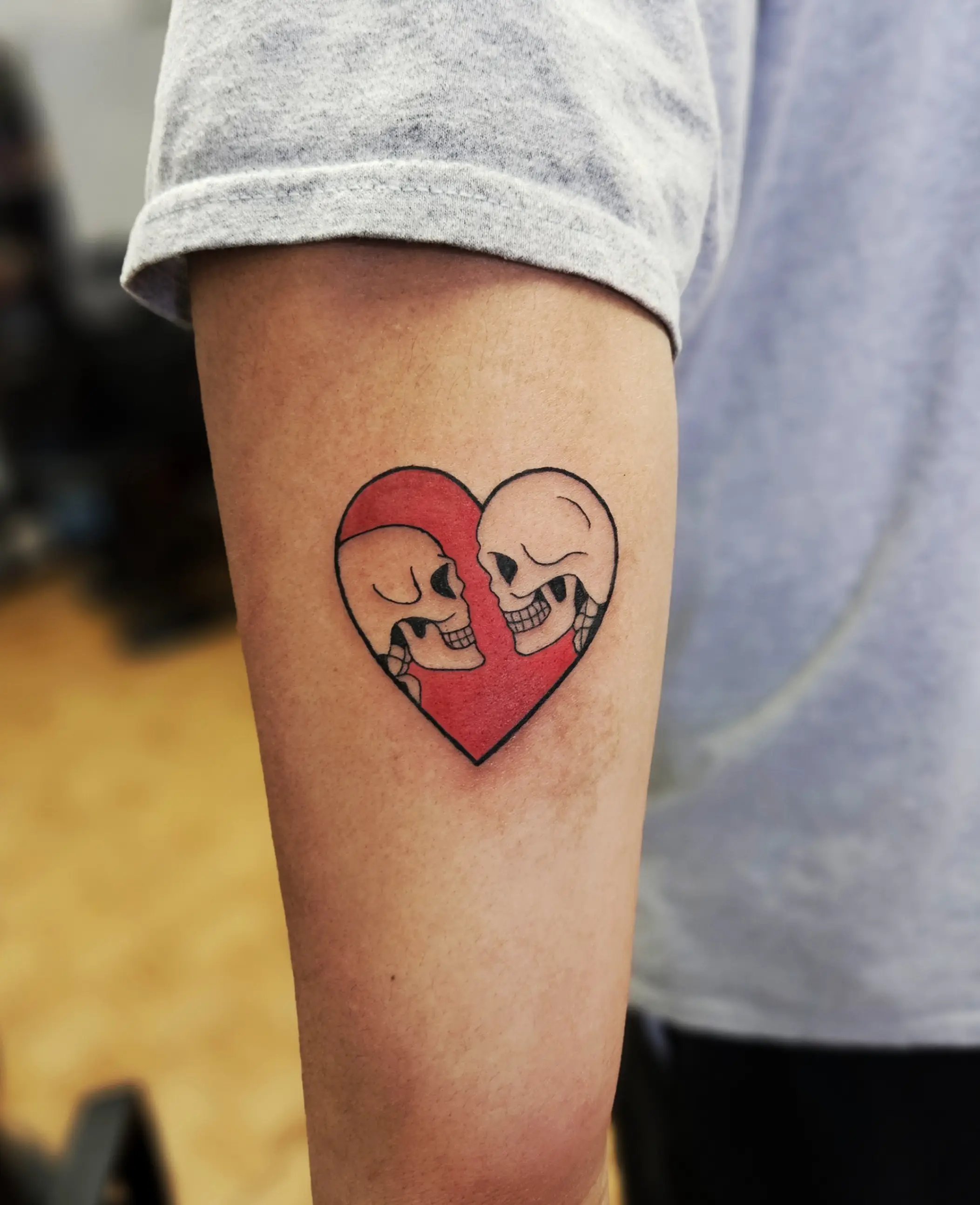 Two skulls forming a heart shape tattoo with bold red background, done in color neo-traditional style.