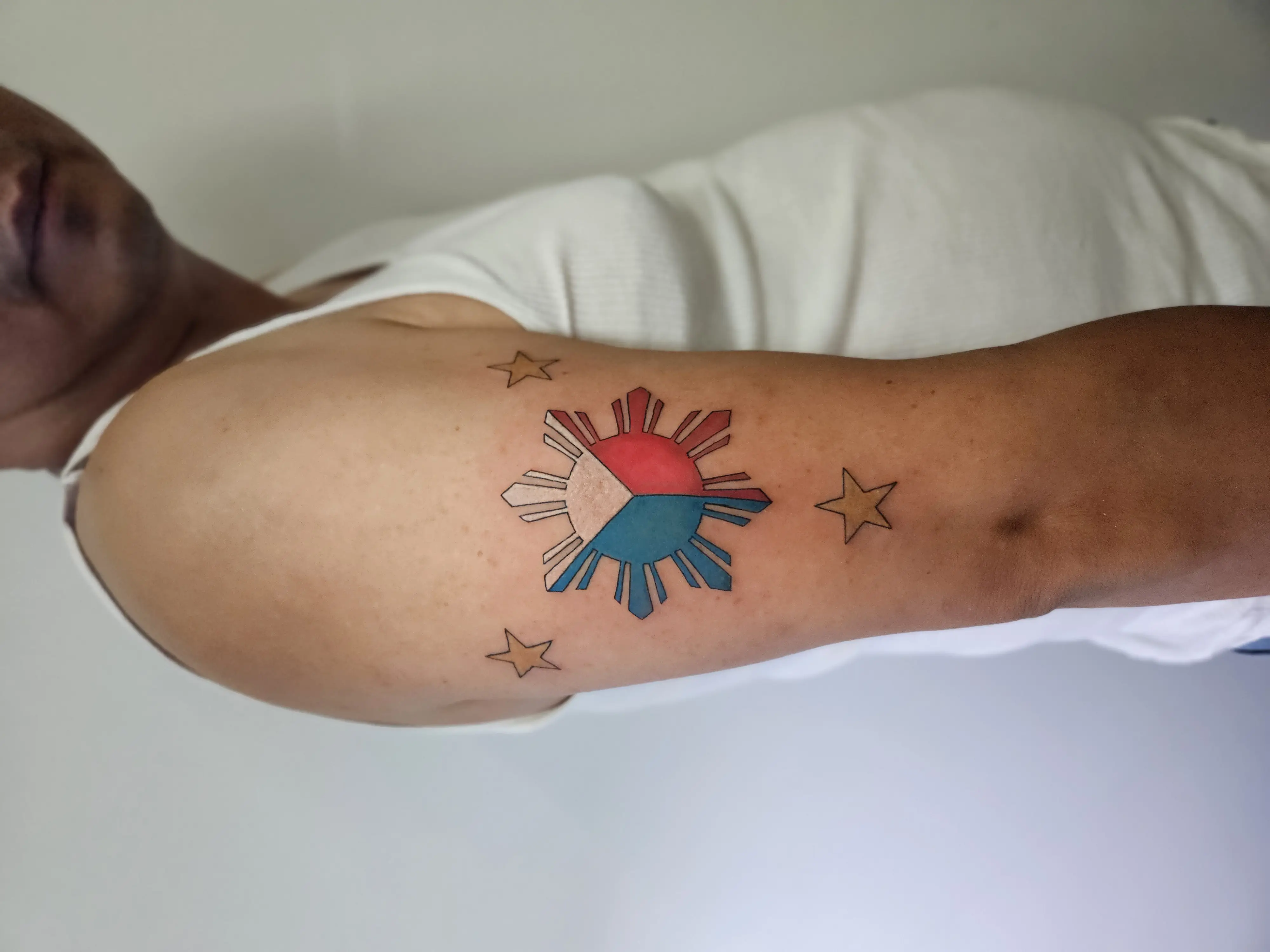Philippine flag-inspired tattoo with sun and three stars in bold color on the upper arm.
