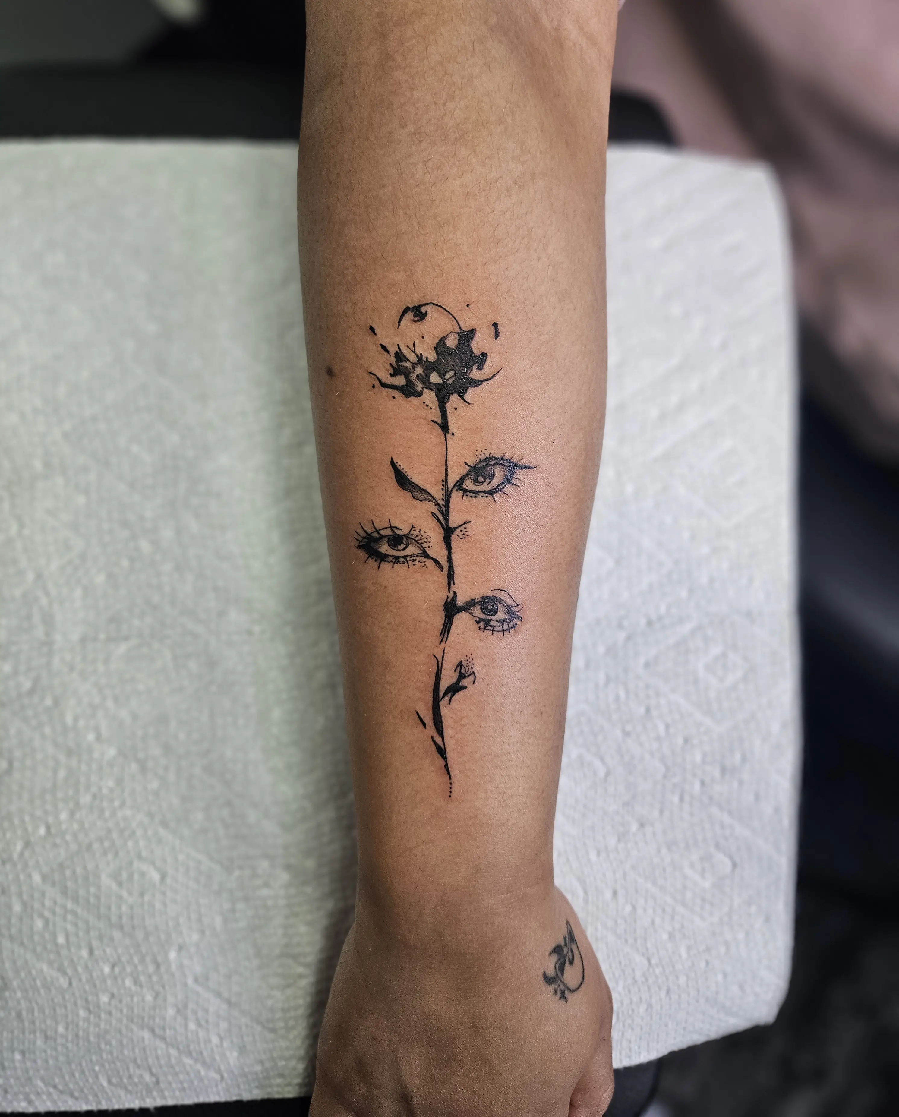 Abstract fine line floral tattoo with eye motifs integrated into the stem design on the forearm.