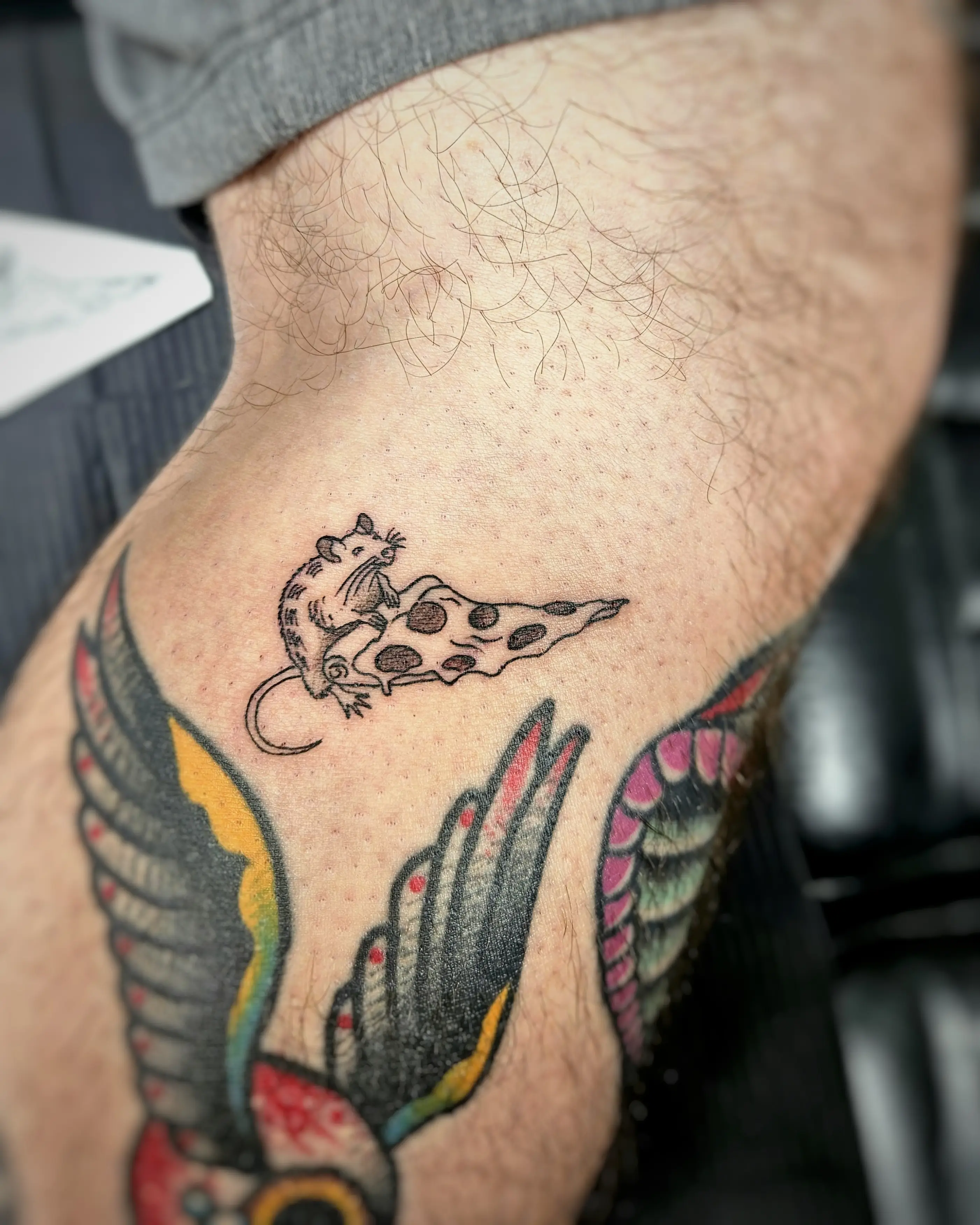 Small, minimalist black outline tattoo of a rat holding a slice of pepperoni pizza, a classic New York City-themed tattoo, placed above a larger traditional wing tattoo.