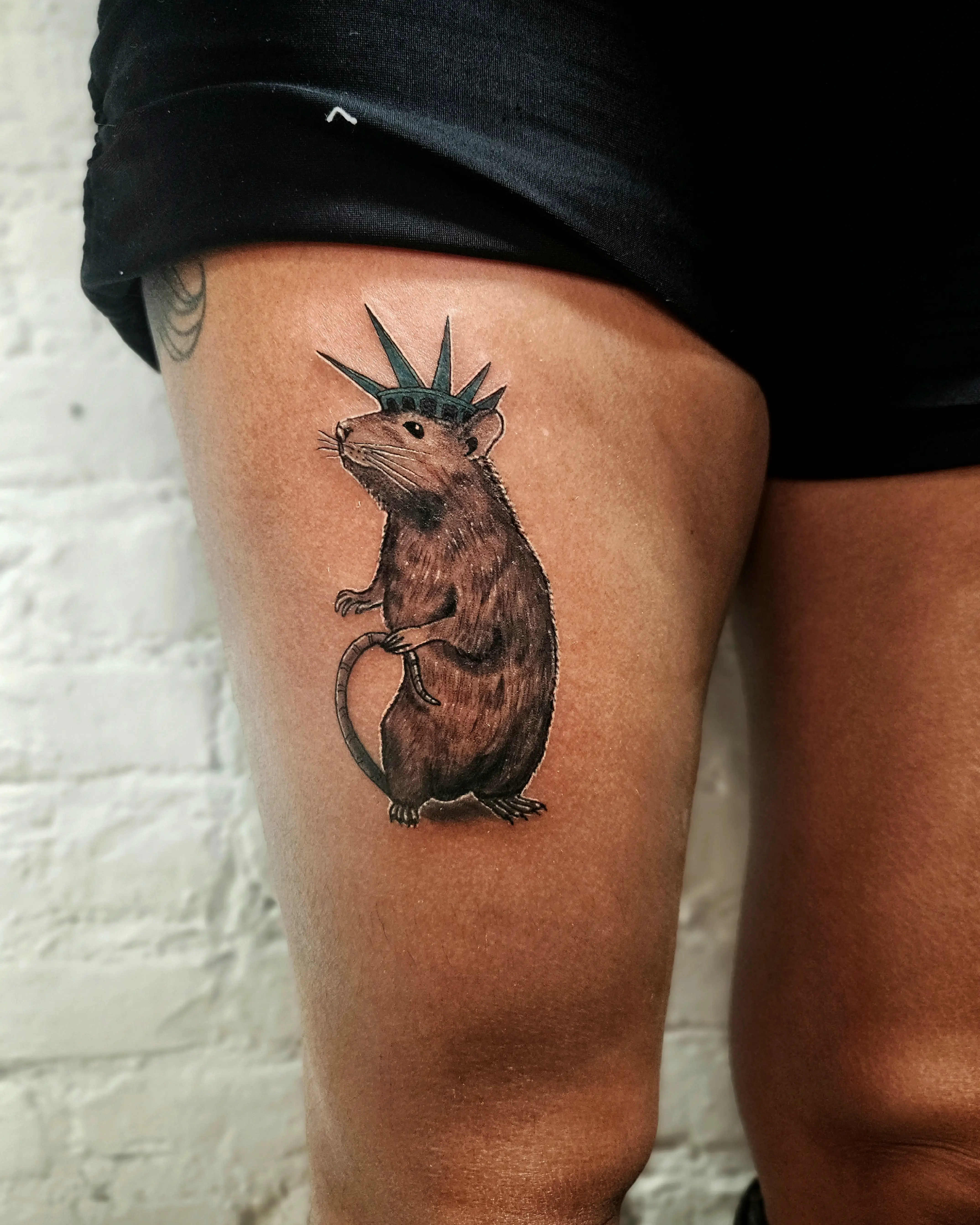 Detailed, illustrative tattoo of a brown rat wearing the crown of the Statue of Liberty, an edgy take on an NYC theme, placed on the upper thigh.