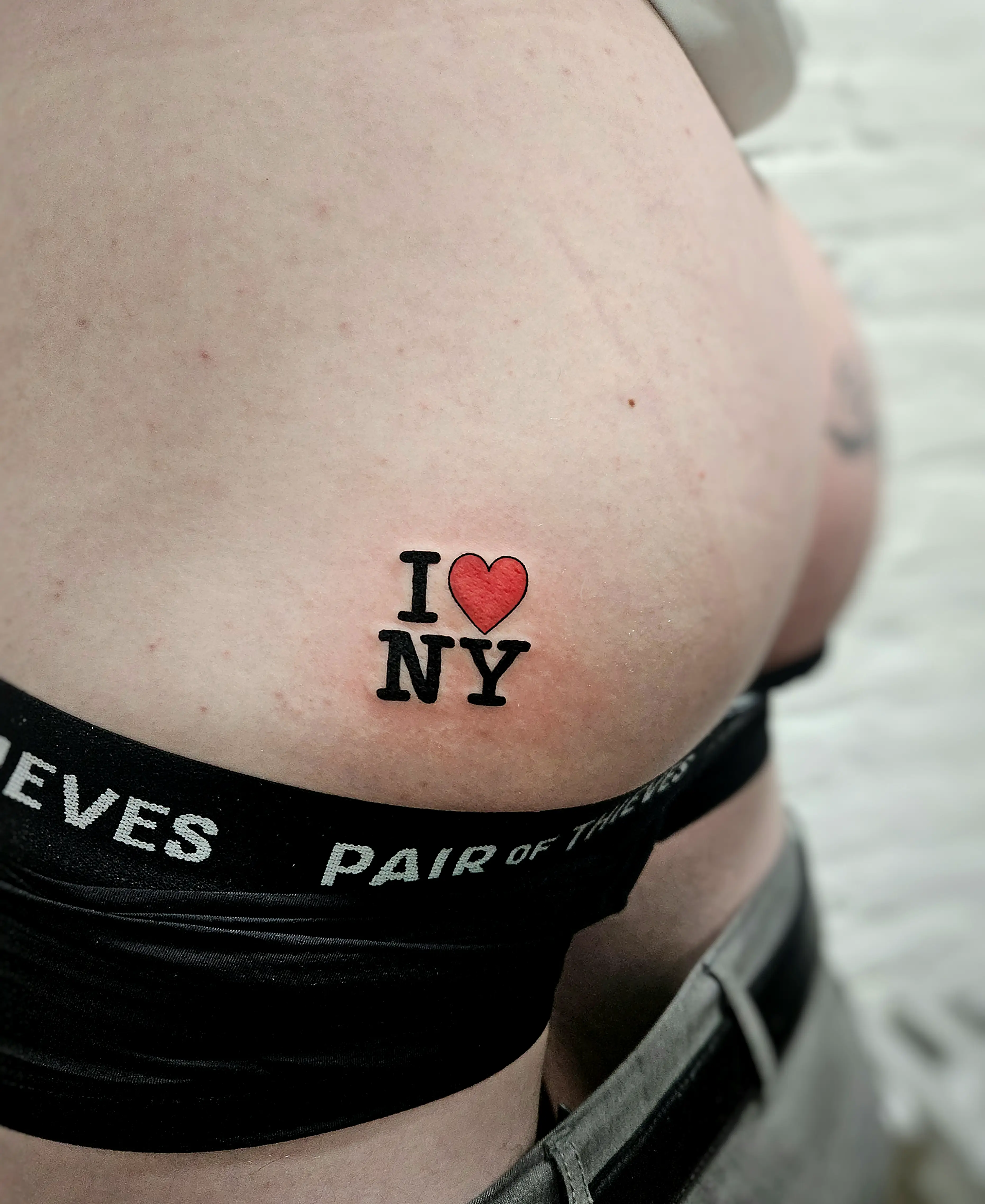 Small, bold tattoo of the classic 'I ❤️ NY' logo with a red heart, placed near the hip/lower back area.